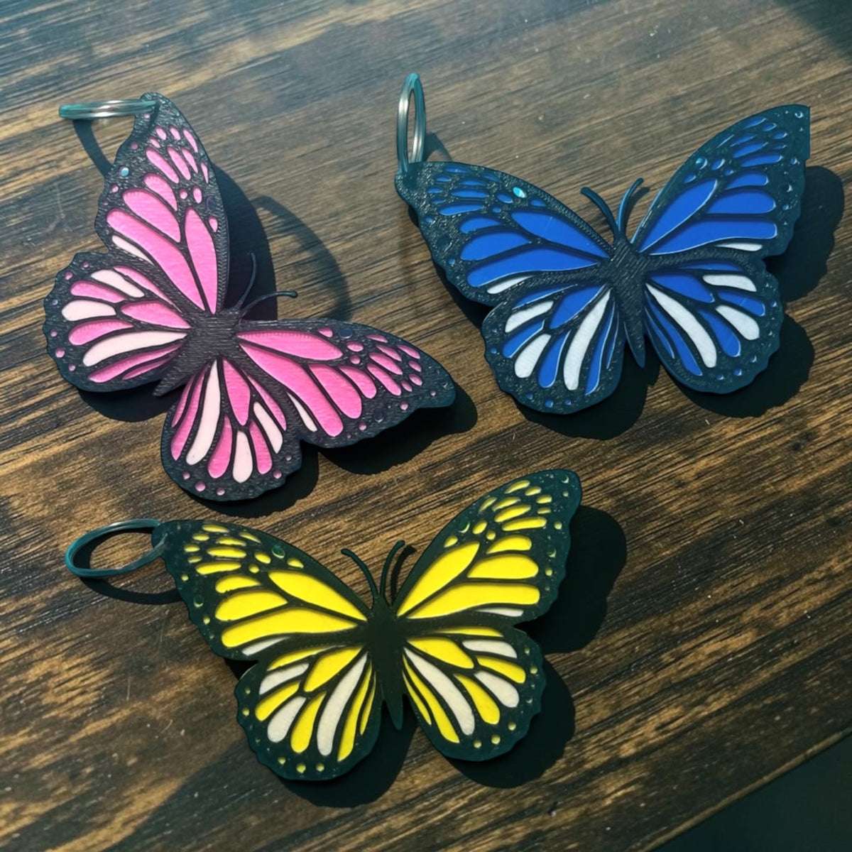 3D printed butterfly keyring set of 3 - pink, blue and yellow on wooden surface
